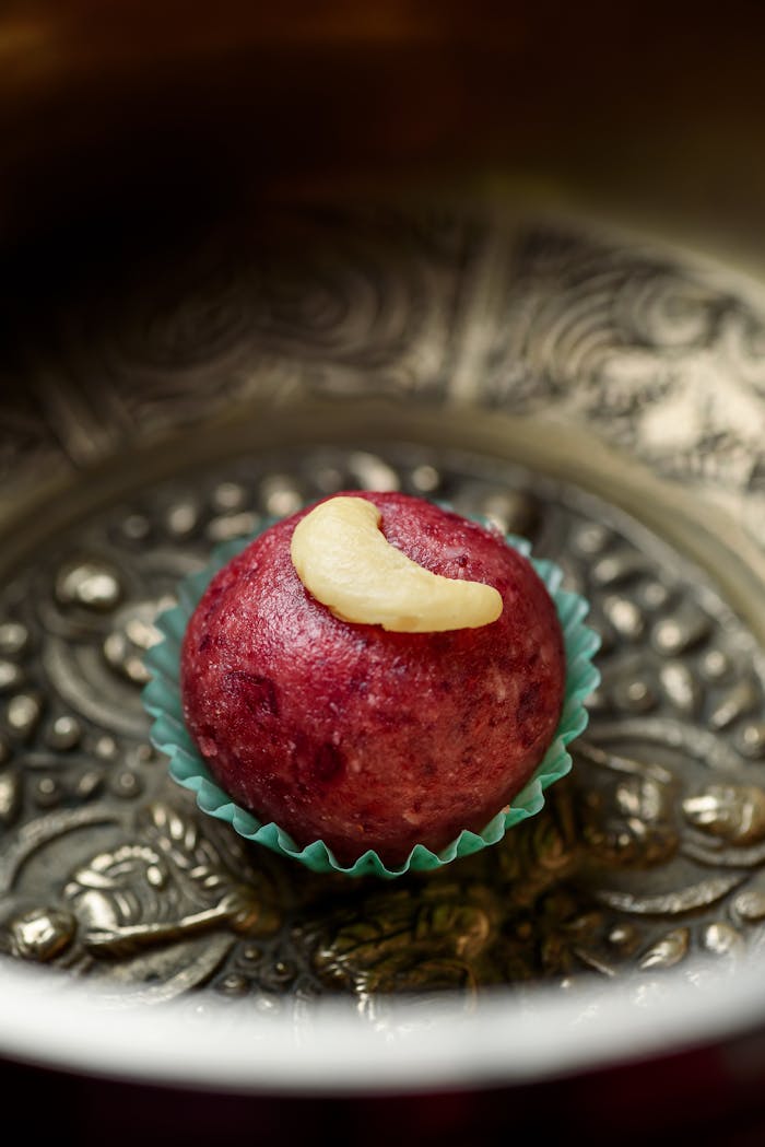 heros-img Close-up of a red laddu with a cashew top, set on an ornate plate.